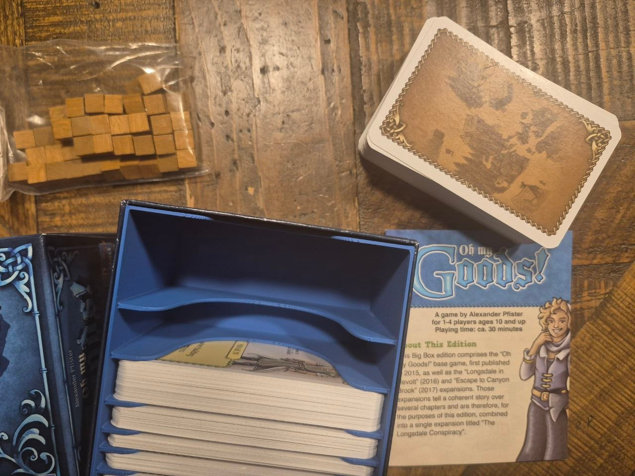 A board game set laid out on a wooden tabletop, including an open blue box with card sleeves, a deck of cards on its illustrated card backing, a bag of small wooden tokens, and part of the game instructions booklet.