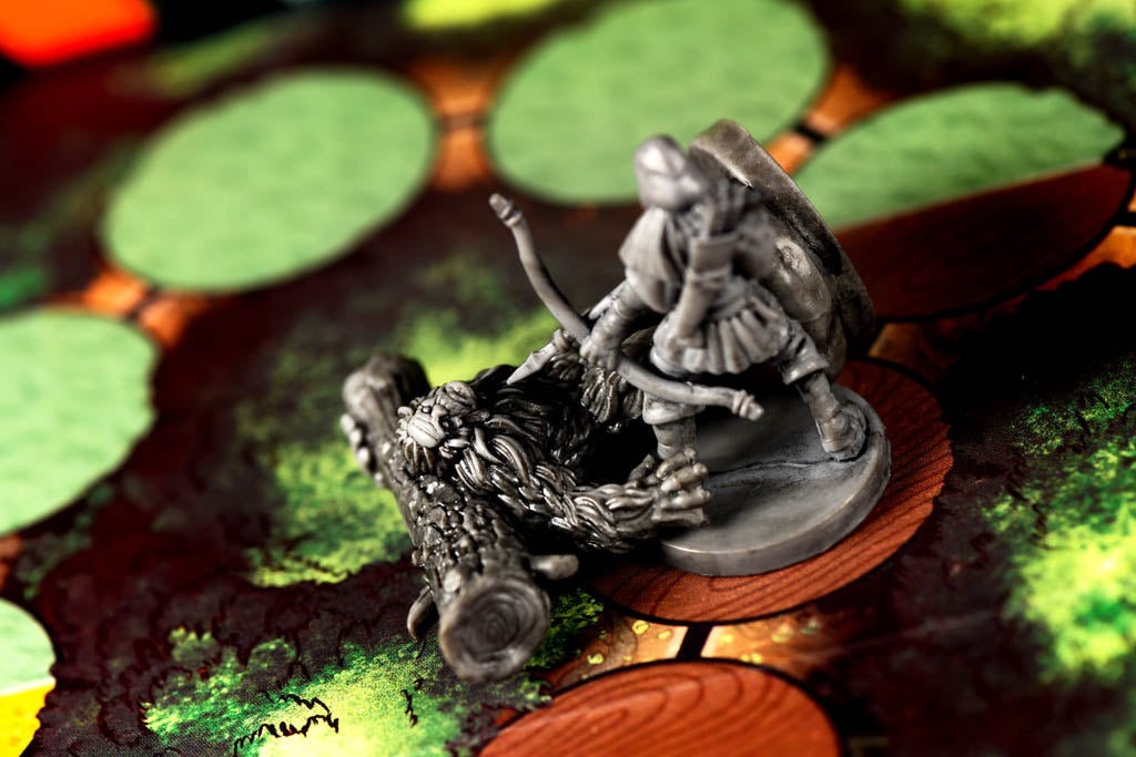 A close-up of a gray fantasy tabletop gaming miniature, depicting a warrior on a log, placed on a printed game board with green and brown terrain details.