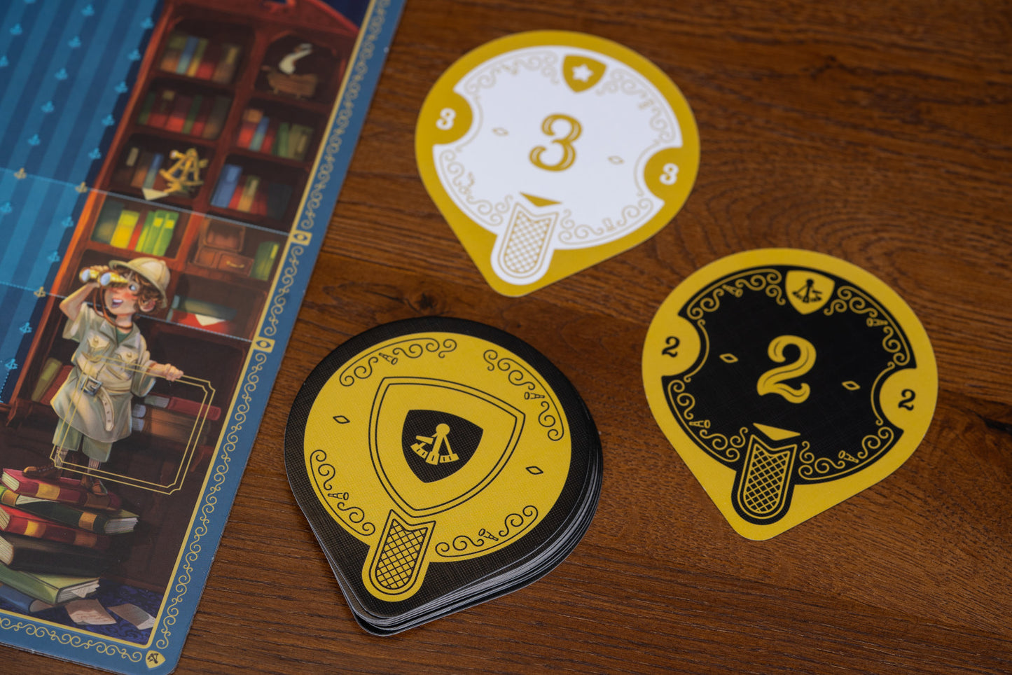 Three decorative cards from Art Society with numbers and symbols on a wooden surface next to a board game box.