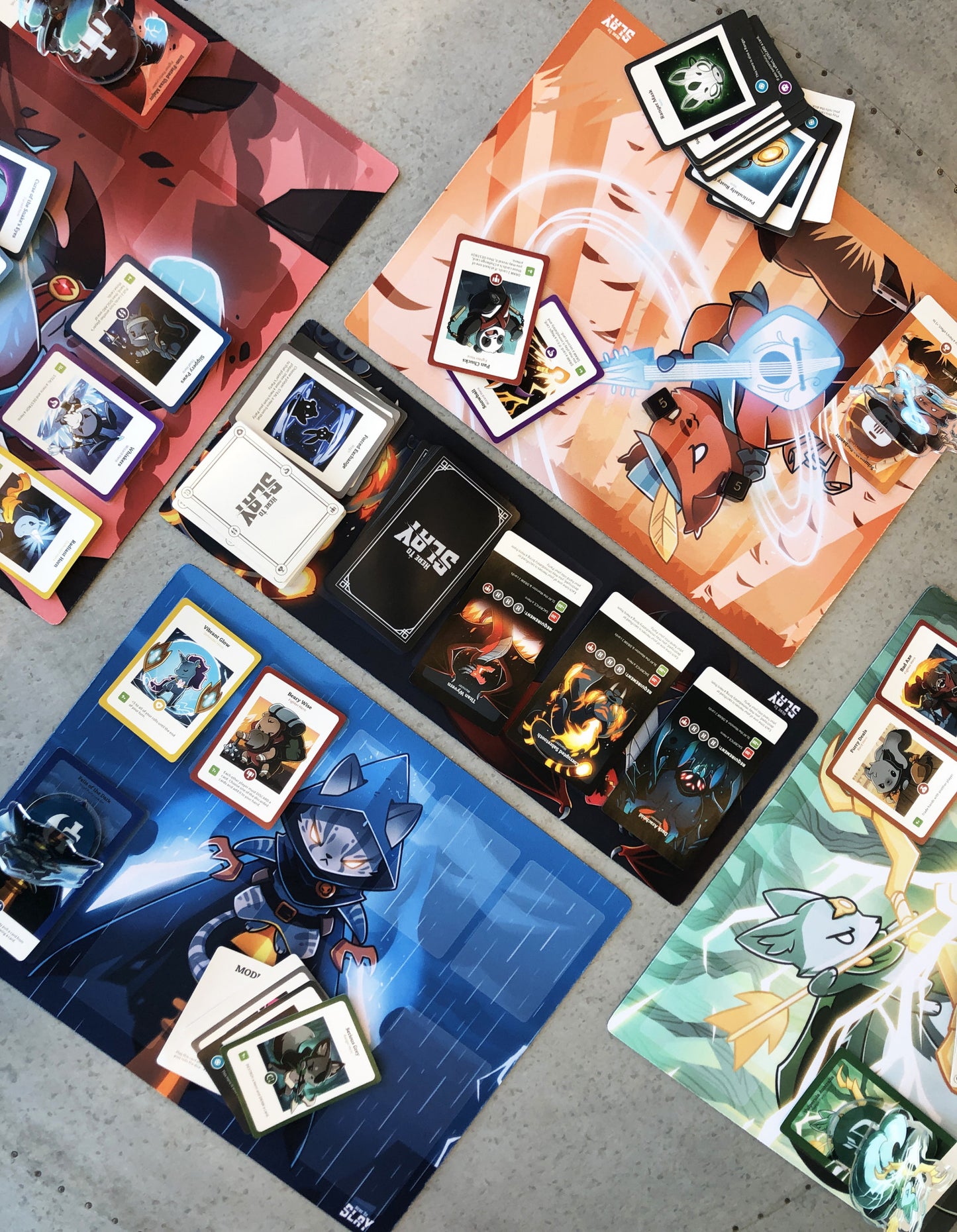 Here to slay board game Collection of trading cards on colorful playmats with character illustrations.