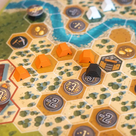 Close-up of a board game board with hexagonal tiles, orange terrain pieces, dark gray resource tokens, and a black player piece on a tabletop.