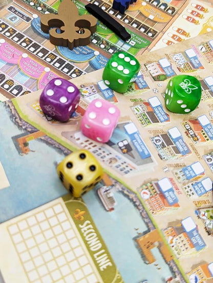 A close-up of a board game setup showing colorful game dice scattered across a detailed city-themed game board. The visible dice include purple, pink, green, yellow, and blue, with a wooden token piece near the top.