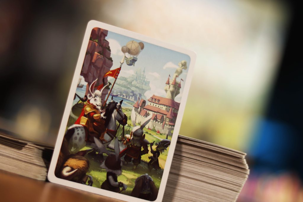 Bunny Kingdom Card with a fantasy scene featuring a character on a horse, surrounded by buildings and trees, with a stack of cards in the background.