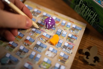 A close-up of a board game in progress on a wooden table, with a hand holding a pencil over a detailed game board, a purple die showing five dots, and a small yellow piece. The box for the game is partially visible in the background.