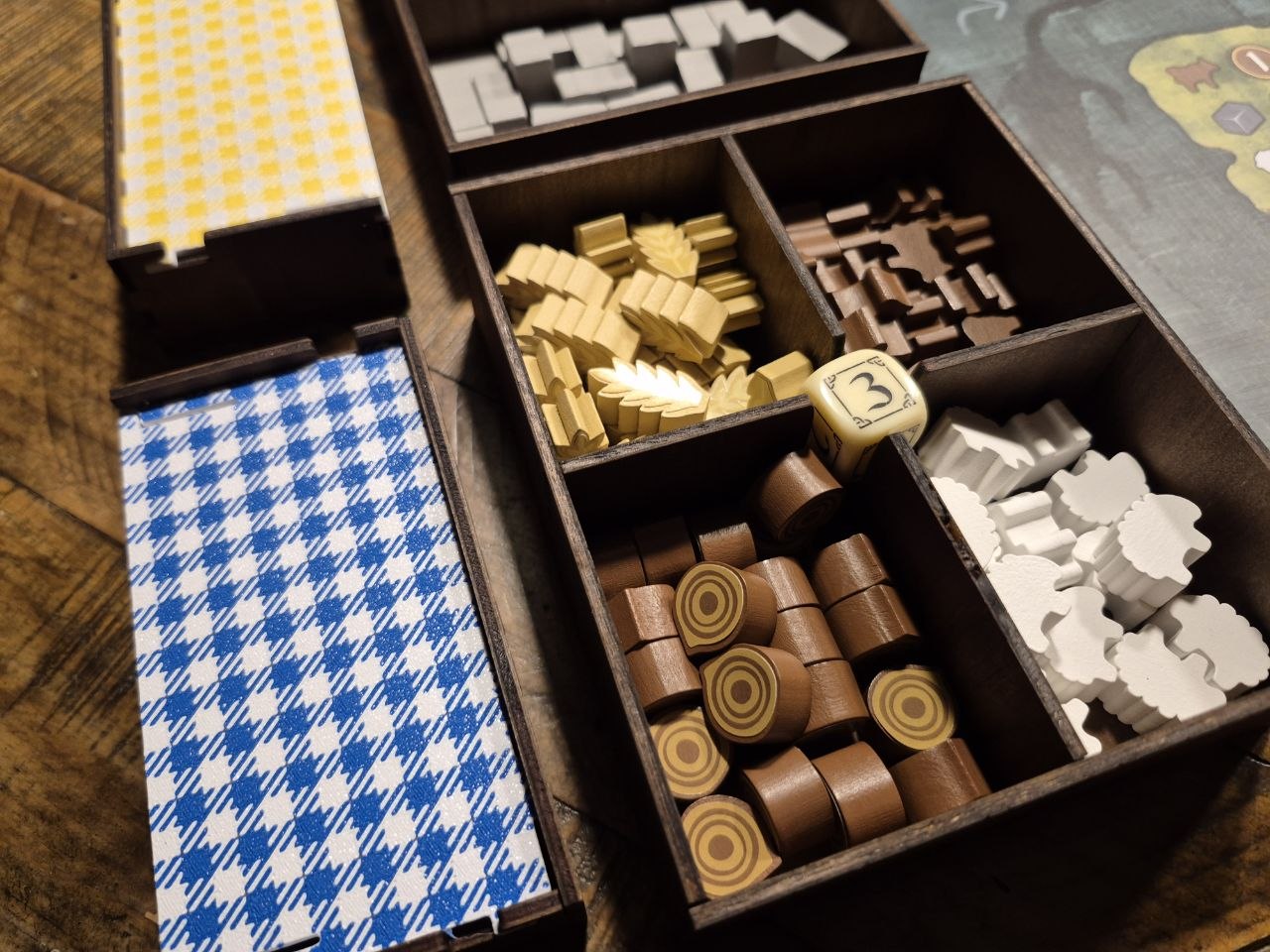A close-up of a wooden divided game box containing various colored game pieces, including gold, brown, white, and blue-and-white patterned components, arranged on a wooden tabletop.