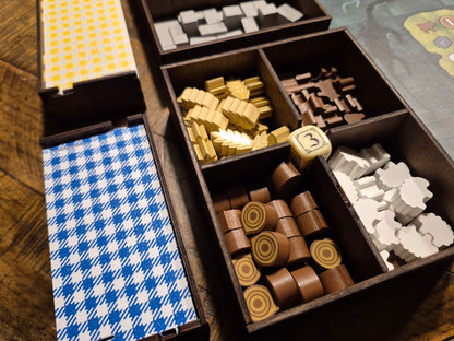 A close-up of a wooden divided game box containing various colored game pieces, including gold, brown, white, and blue-and-white patterned components, arranged on a wooden tabletop.