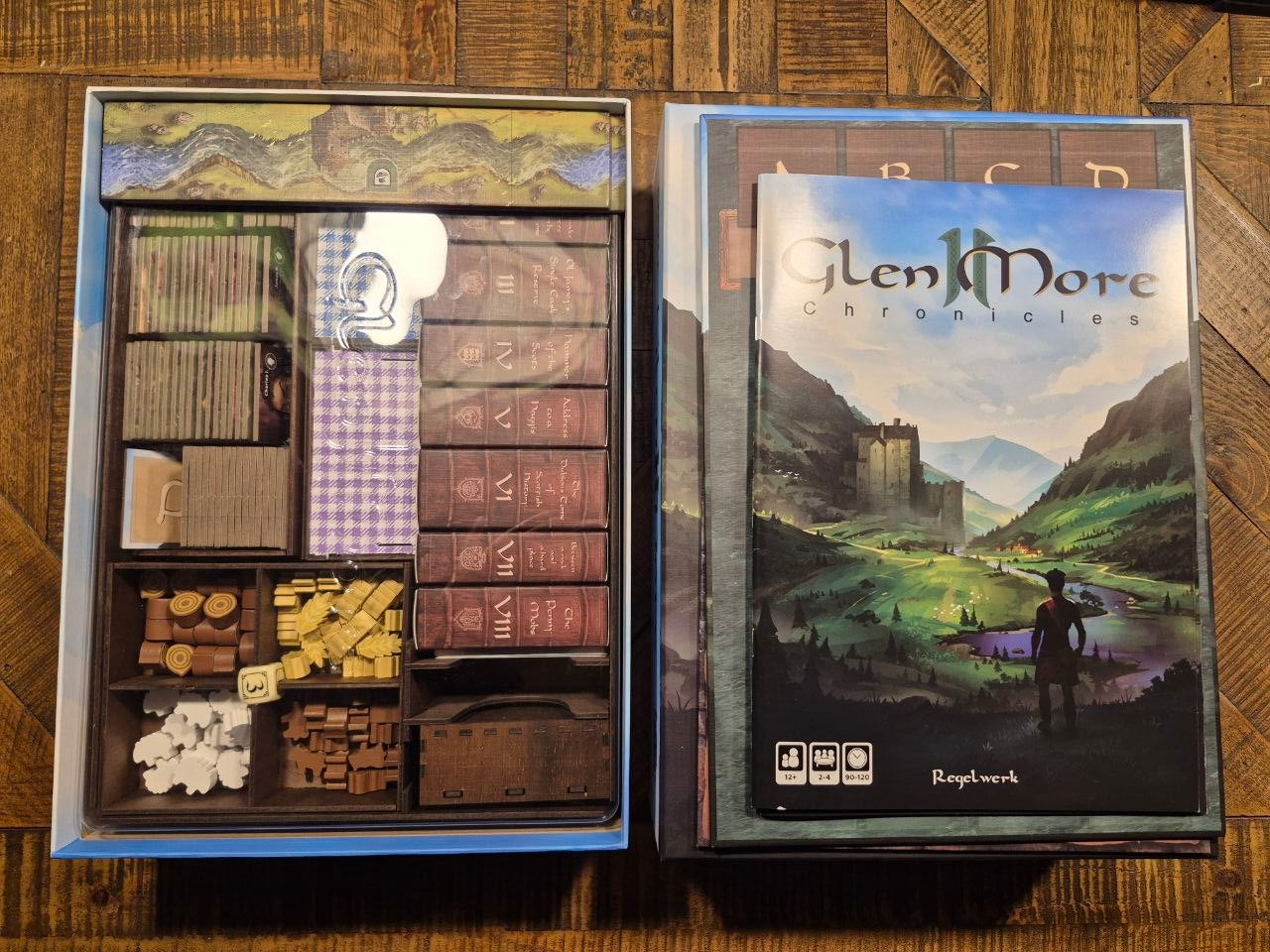 Glen More: Chronicles II board game oragniser shown open, displaying its various components including rulebooks, play money, tokens, and cards, alongside the illustrated game box on a wooden surface.