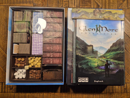 The box set for the board game Glen More: Chronicles, shown open to reveal the organized contents including rulebooks, cards, and tokens, placed on a wooden table.