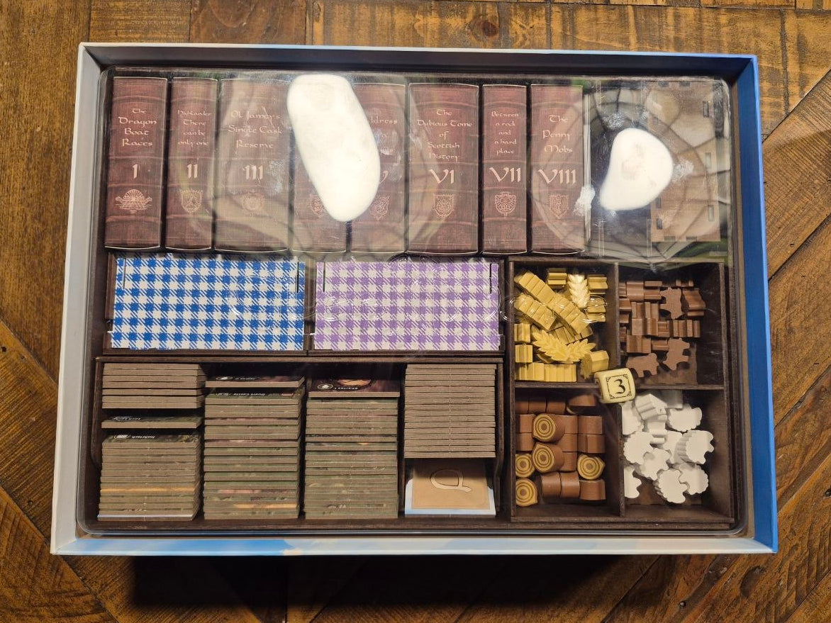 An open boxed game set displayed on a wooden surface, showing organized compartments with miniature cards, tokens, blocks, and small game pieces under clear plastic cover.