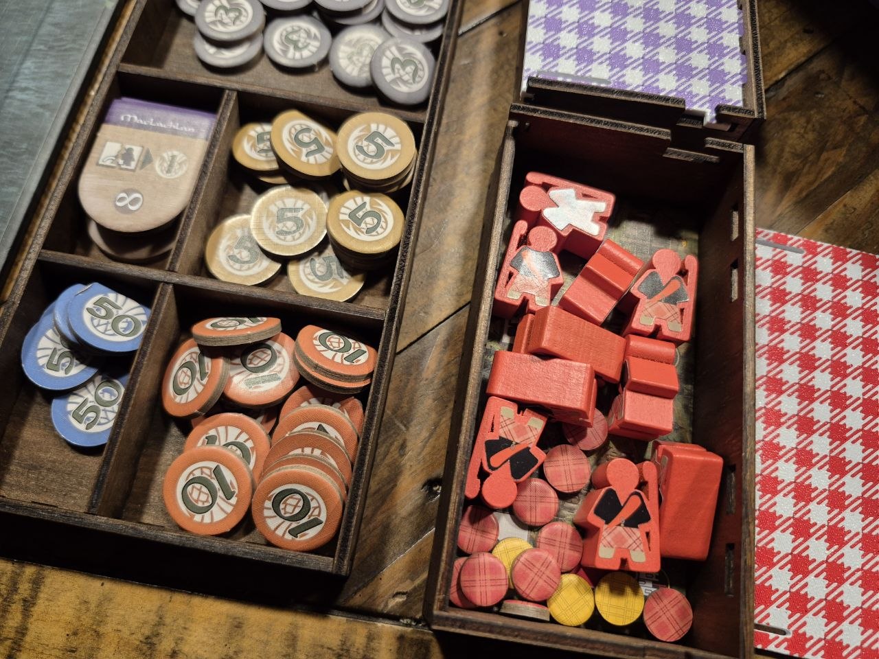 Open wooden game boxes filled with round and shaped tokens and red player pieces, photographed on a wooden tabletop. A red-and-white checkered card is visible at the right edge.