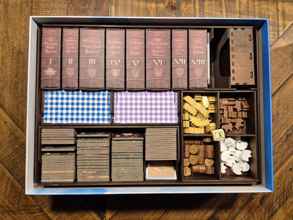 An overhead view of a boxed board game set arranged in a deluxe timber tray. The set includes brown book-like pieces, blue-and-white checkered cards, purple-and-white checkered cards, gray and brown token pieces, and white paper pieces, all on a wooden surface.