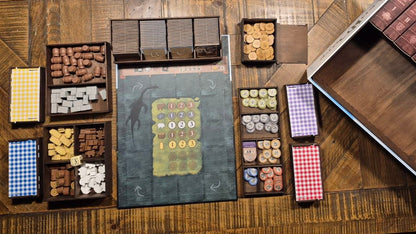 Overhead view of a wooden tabletop filled with boxed and tray-based craft or game components, including small blocks, chips, cards, and tools like orange-handled scissors and a pink mug.
