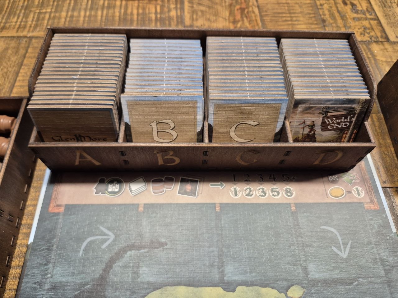 Glenmore wooden game box filled with tile decks arranged in four columns labeled A, B, C, and D, placed on top of a board game mat on a wooden tabletop.