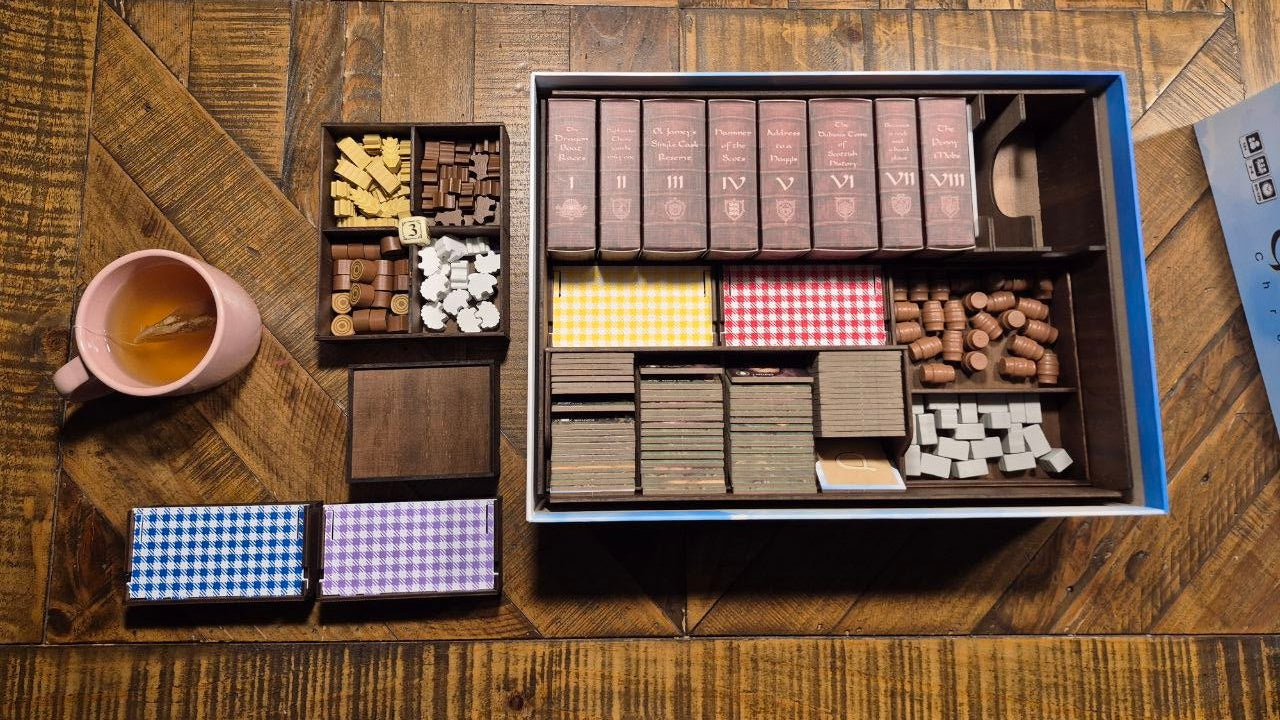 An overhead view of a wooden game board or card game set organized in a blue storage box, placed on a rustic wooden table. The set includes multiple compartments filled with cards, tokens, and small game pieces in various colors.