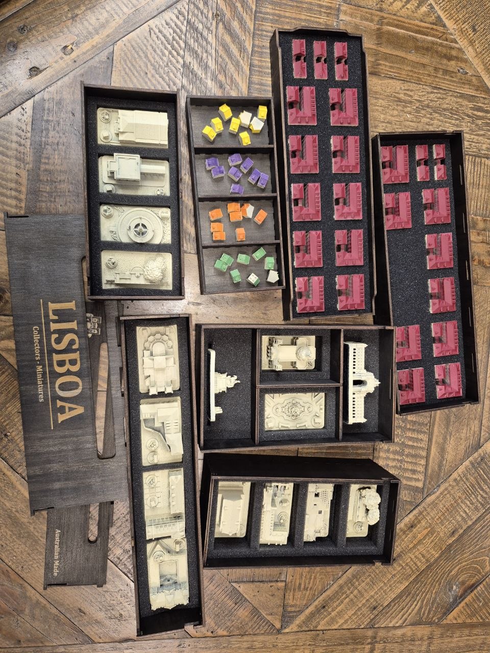 A set of miniature model train tracks and terrain pieces from Games Workshop's 'Lisboa - Portugal Expansion Set', displayed in their black foam trays and wooden box on a rustic wooden floor.