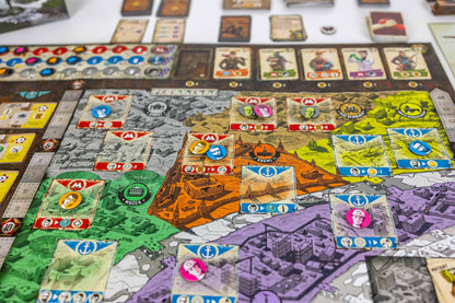 Resurgence colorful board game map with cards, tokens, and game pieces arranged on a tabletop. The map features distinct colored regions in green, orange, yellow, purple, and gray, with various illustrated cards and circular player tokens spread across the surface.