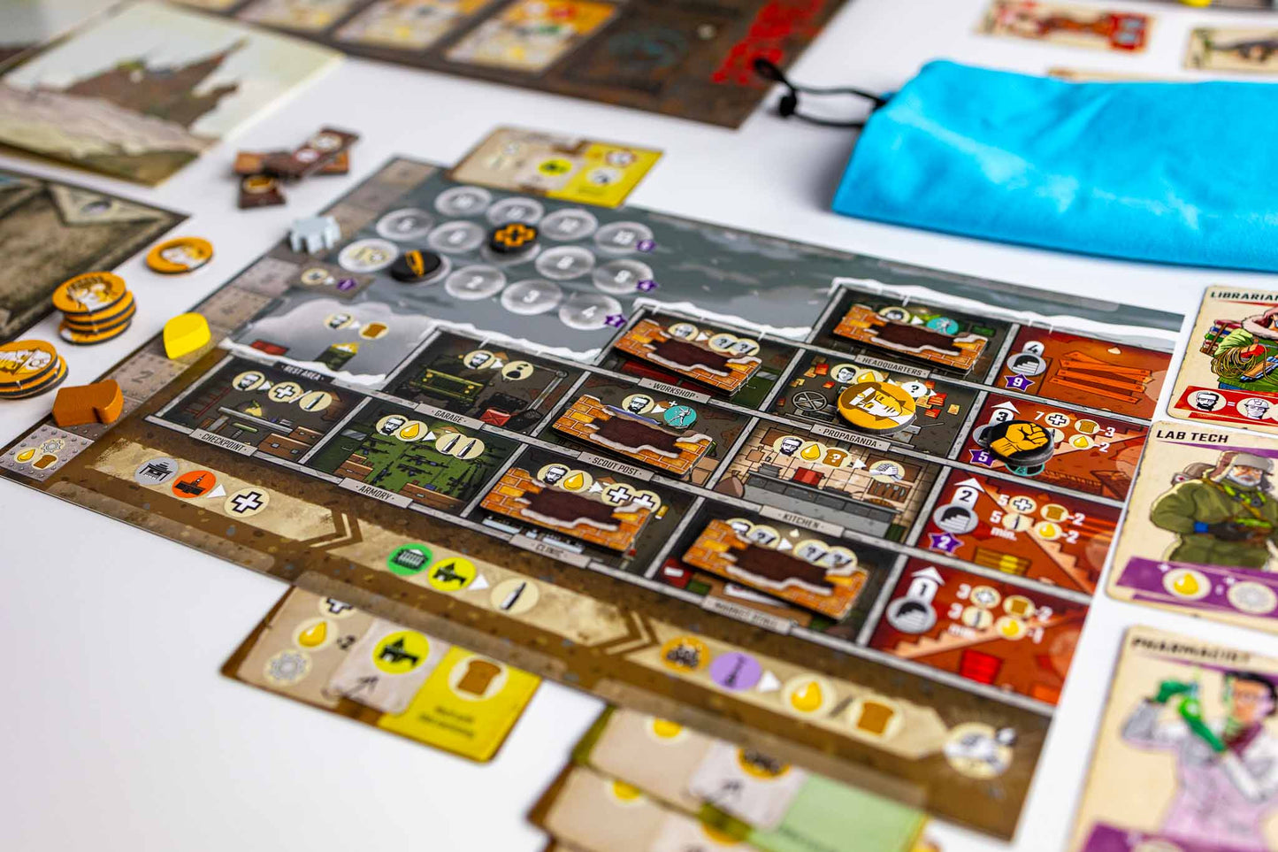 Resurgence board game setup on a white surface, showing a detailed game board with cards, tokens, coins, and a bright blue drawstring pouch.
