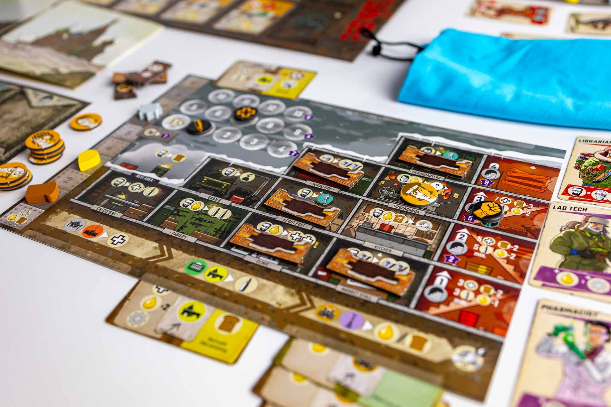 Resurgence board game setup on a white surface, showing a detailed game board with cards, tokens, coins, and a bright blue drawstring pouch.