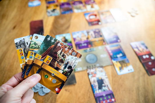 Hand holding a collection of board game cards on a wooden table with more cards scattered around.