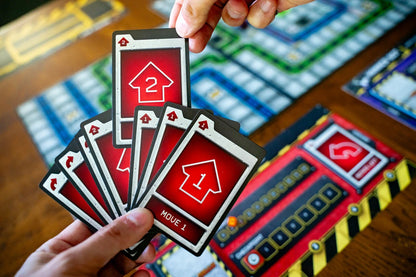 Hand holding game cards Robo Rally  with red and black design, likely from a board game, with a blurred game board in the background.