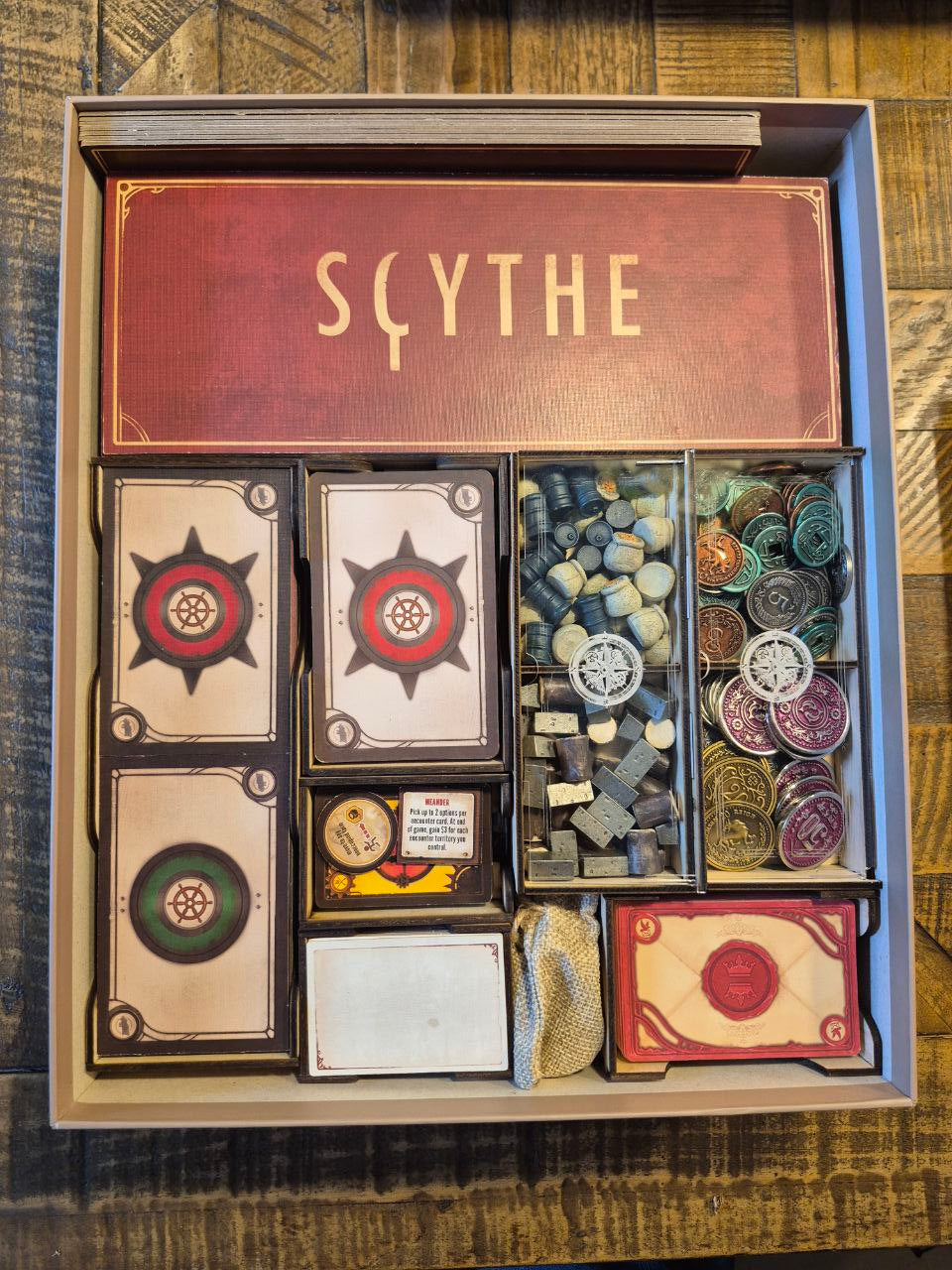 Board game components including cards and pieces in a 'Sythe' box on a wooden surface.