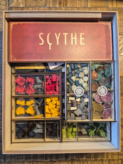 Board game components in a box labeled 'Scythe' on a wooden surface