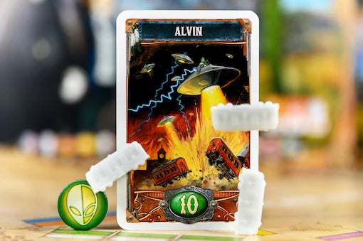 A close-up of a card game card titled 'ALVIN' showing a fiery alien spaceship scene, with white plastic standoffs holding it upright on a game board. A green alien token is visible at the lower left, and the background is softly blurred.