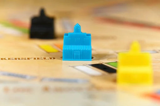 A close-up shot of a blue plastic building game piece standing on a printed board, with other colorful pieces blurred in the background.