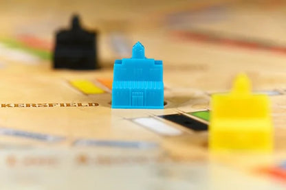 A close-up shot of a blue plastic building game piece standing on a printed board, with other colorful pieces blurred in the background.