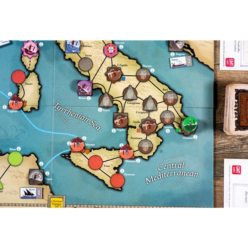 A close-up of a board game map showing the Italian peninsula and surrounding Tyrrhenian and Central Mediterranean Sea, with game pieces, cards, and dice on a wooden tabletop.