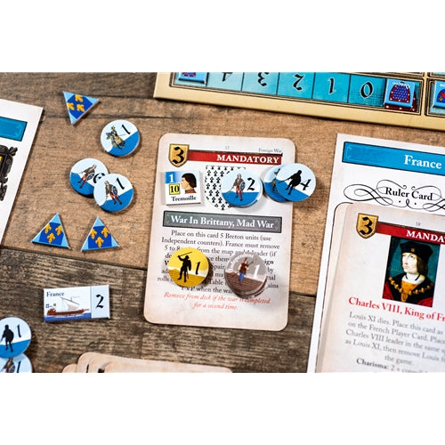Board game components arranged on a wooden tabletop, including cards, blue numbered tiles, round player tokens, triangular terrain pieces, and a ruler-like piece. A central card reads 'War in Brittany, Mad War' and 'MANDATORY,' with a France ruler card partially visible on the right.