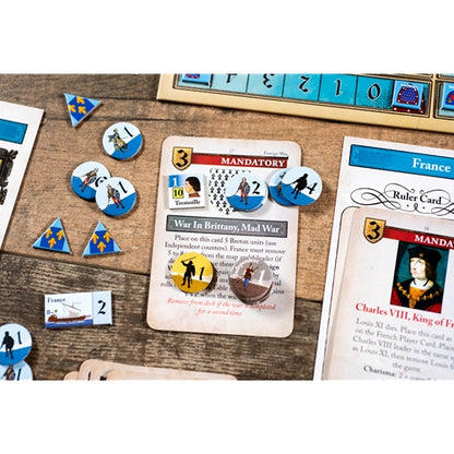 Board game components arranged on a wooden tabletop, including cards, blue numbered tiles, round player tokens, triangular terrain pieces, and a ruler-like piece. A central card reads 'War in Brittany, Mad War' and 'MANDATORY,' with a France ruler card partially visible on the right.
