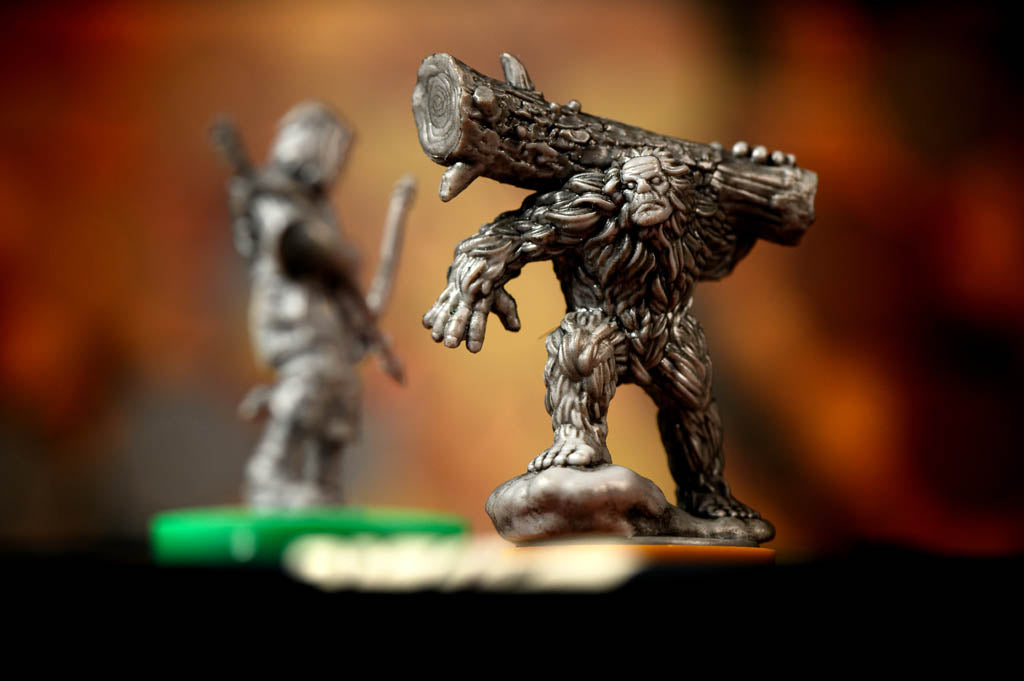 A detailed pewter-style miniature figurine of a Bigfoot-like creature carrying a large log on its back, shown in sharp focus against a blurred warm brown and orange background. A second out-of-focus miniature is visible in the background.