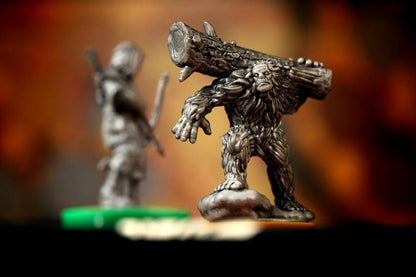A detailed pewter-style miniature figurine of a Bigfoot-like creature carrying a large log on its back, shown in sharp focus against a blurred warm brown and orange background. A second out-of-focus miniature is visible in the background.