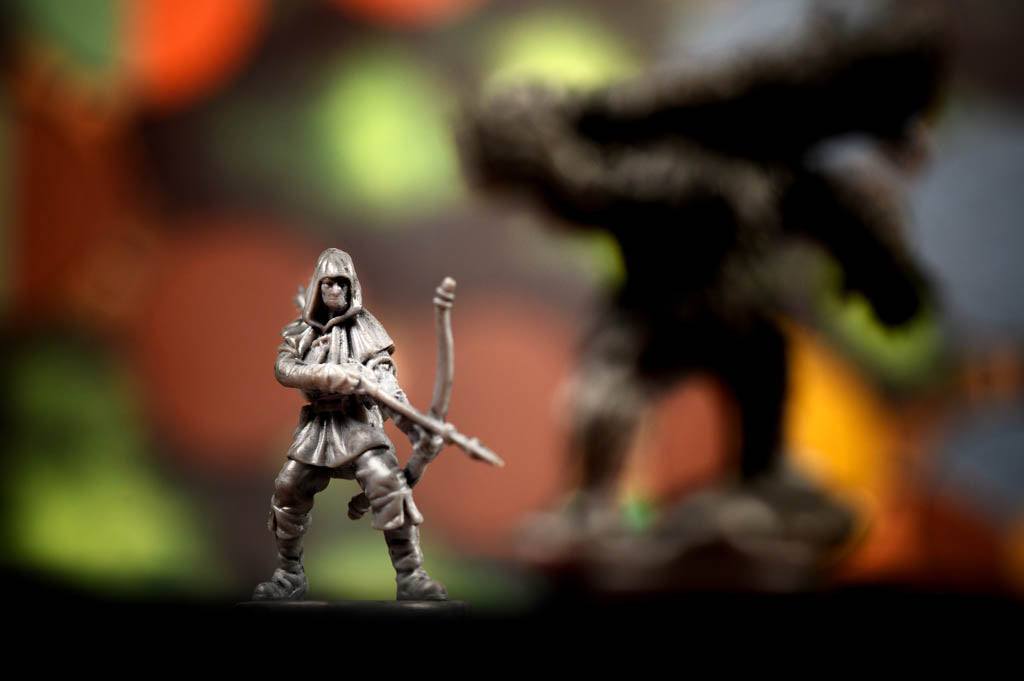 A small unpainted gray plastic fantasy warrior miniature figurine holding a bow, standing in front of a heavily blurred dark fantasy creature silhouette on a colorful, out-of-focus background.