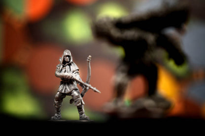 A small unpainted gray plastic fantasy warrior miniature figurine holding a bow, standing in front of a heavily blurred dark fantasy creature silhouette on a colorful, out-of-focus background.