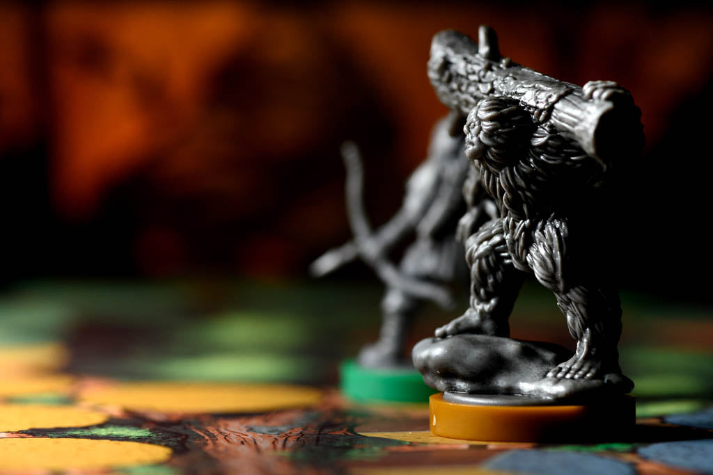 A close-up of a dark metallic fantasy creature figurine standing on a round base, with another blurred figurine behind it on a colorful tabletop surface.