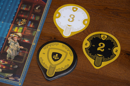 Three decorative cards from Art Society with numbers and symbols on a wooden surface next to a board game box.