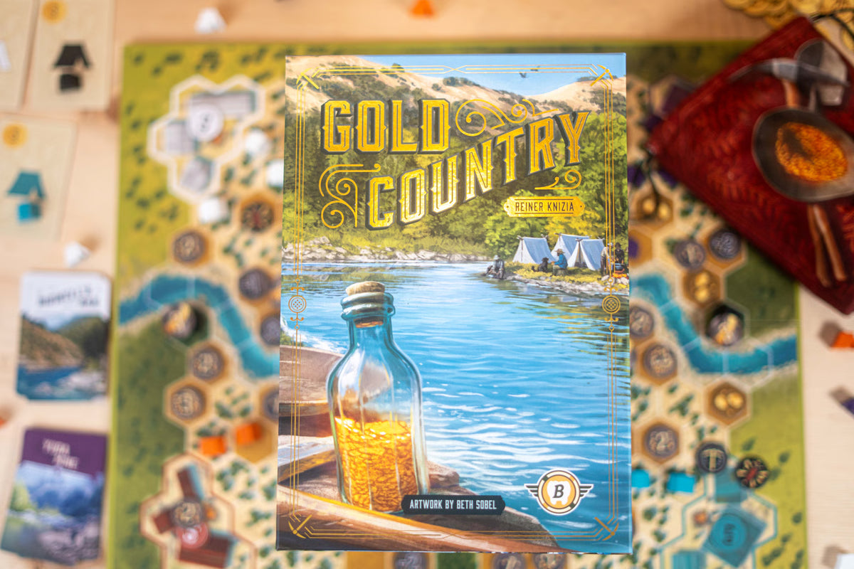 Board game box for Gold Country: Single Rides, shown standing upright on a game board with cards and tokens around it. The box art features a scenic lake and mountains with a bottle of gold in the foreground.