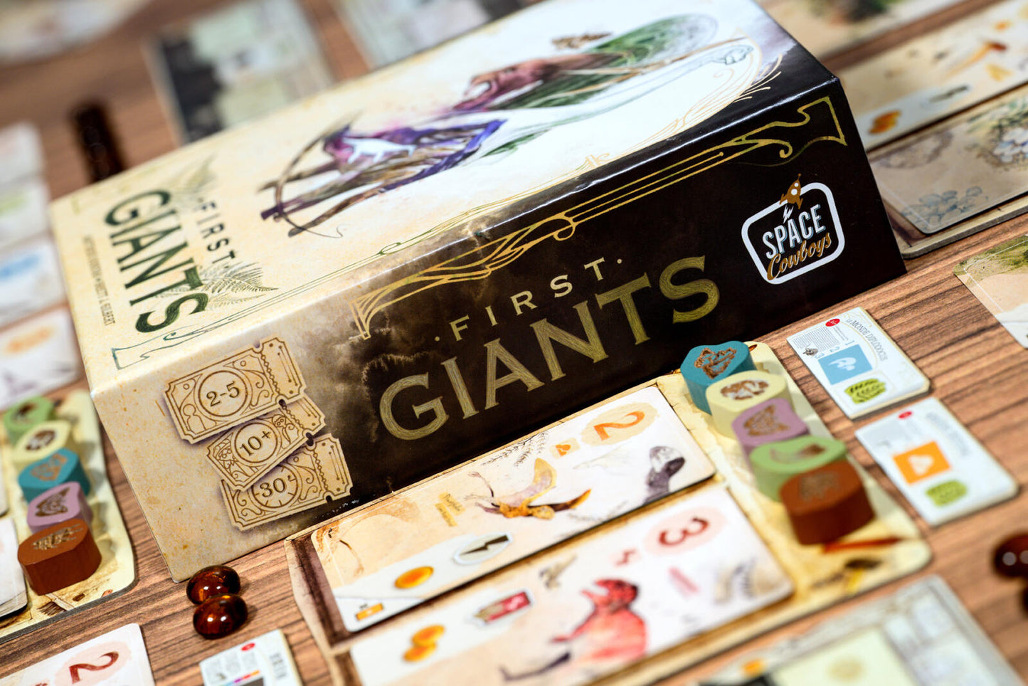 Board game box for 'First Giants' by Space Cowboys on a wooden table with game pieces.