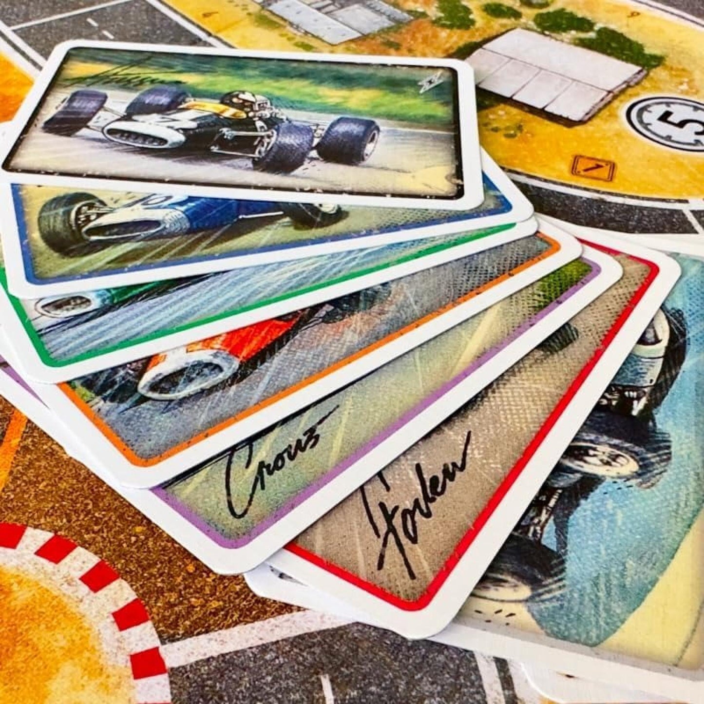 A close-up of several racing-themed cards fanned out over a game board. The cards feature vintage race car illustrations, white borders, and signatures, with a colorful tabletop game surface visible underneath.