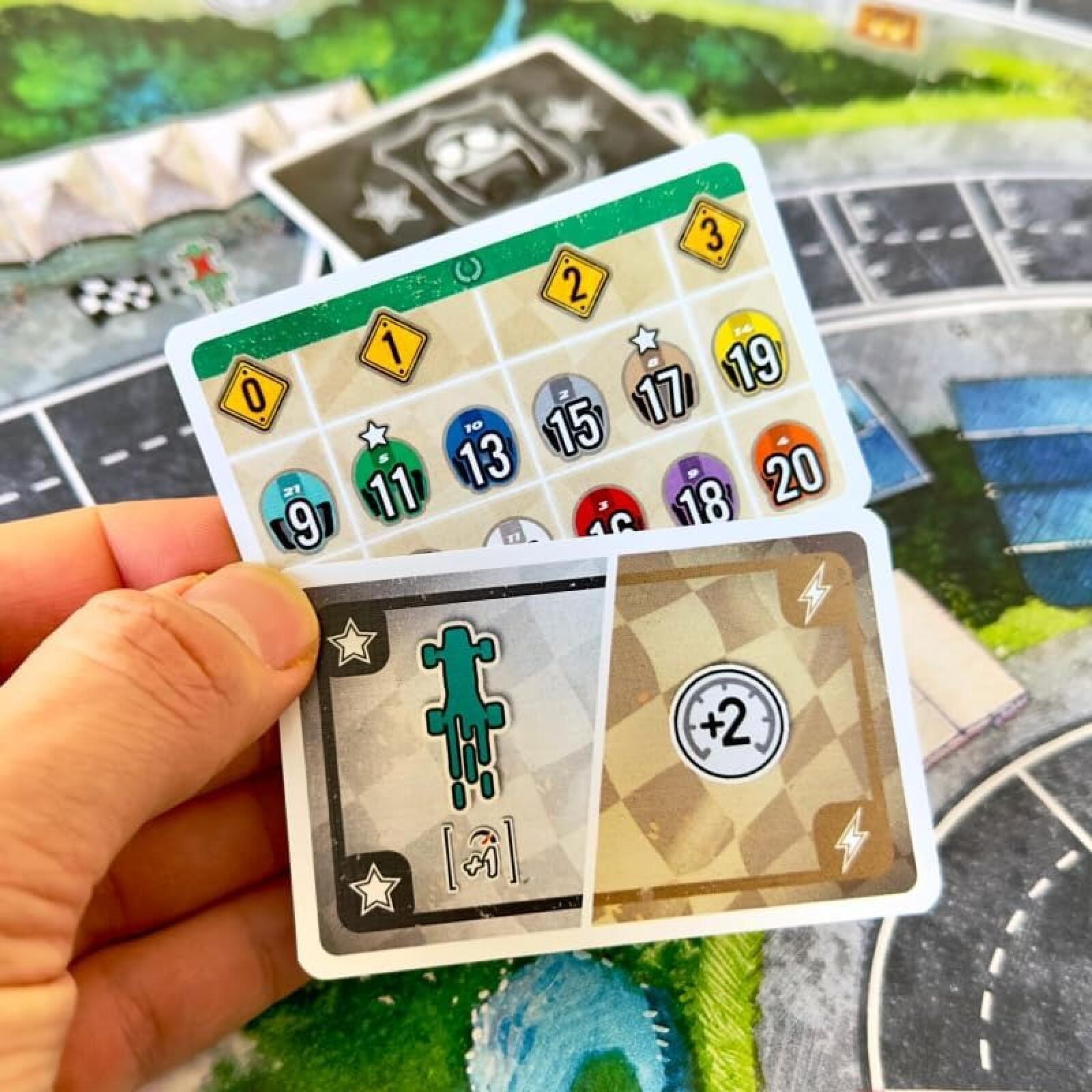 A hand holding two cards from the board game Catan, showing resource and action cards over a green and gray game board.