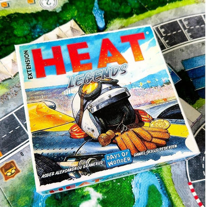 The board game box for 'Heat Legends: Days of Wonder' expansion is shown at an angle, resting on a green and blue illustrated game board. The box features a colorful illustration of a race car driver in a helmet with a yellow sun visor, and the title 'HEAT LEGENDS' in large red and white letters.