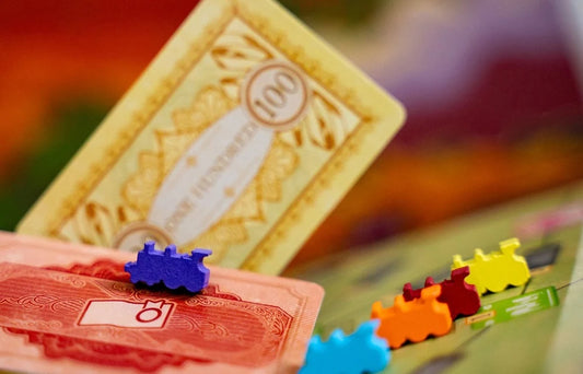 A close-up of a board game setup with colorful toy train pieces on orange and yellow game cards, photographed on a blurred multicolor surface.