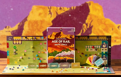 The board game 'Age of Rail: South Africa' displayed on a tabletop with its main box in the center, flanked by the green and blue game boards, cards, and tokens. The background is a stylized purple and yellow mountain graphic.