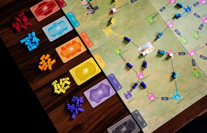 A close-up of a board game in progress on a wooden table, showing a green game board with many colorful cards, tokens, and small figurines arranged in a complex layout.