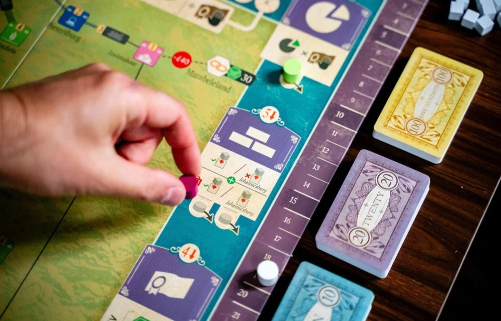 A close-up of a hand playing a board game, placing a pink token on a colorful game board. Several card decks are stacked on a dark wooden table.