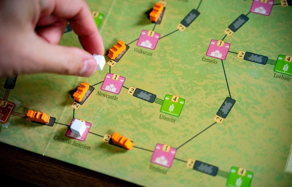 A close-up of a hand placing a small white token onto a green board game map with colorful route tiles and orange train pieces.