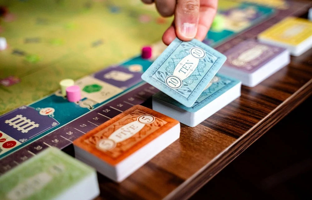 A close-up of a hand placing a blue playing card onto a board game table, with other colorful cards and a game board visible in a real tabletop setting.