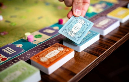 A close-up of a hand placing a blue playing card onto a board game table, with other colorful cards and a game board visible in a real tabletop setting.